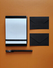 Black and white empty notebook, pen, black envelopes on orange background, top view.
