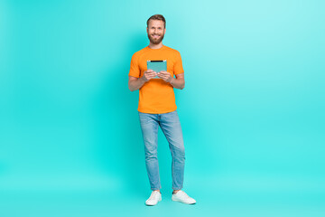 Full size photo of optimistic handsome cheerful man dressed orange t-shirt white sneakers hold tablet isolated on teal color background