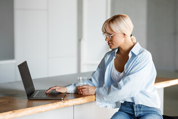 A young blond girl is working on a laptop at home in the kitchen. There is natural lighting, she is wearing a white T-shirt and a blue shirt. Freelance, modern life, business.