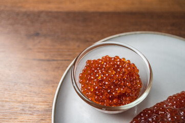 Fresh raw salmon roe eggs sag Ikura or sujiko, in a glass bowl and plate on wooden table top. A popular Japanese food ingredient for making sushi. 