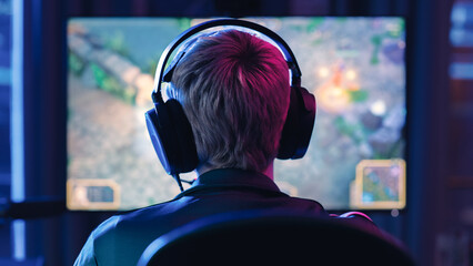 Back of the Head Angle of a Young Female Gamer Wearing Headset and Playing a Video Game on Personal Computer in a Neon Lit Living Room at Home. Cozy Evening for a Pro PLayer at Home in Loft Apartment. © Gorodenkoff