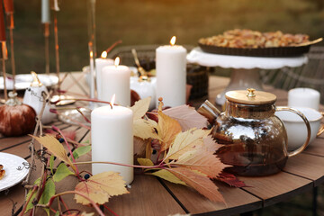 Cozy autumn picnic in the park. Close up of glass kettle with hot black tea, classic cups, homemade apple pie, maple leaves and burning candles on wooden table outdoors.