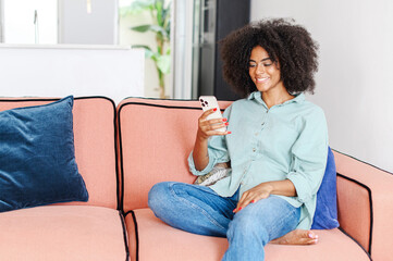 Portrait of smiling woman sitting on sofa at home, using mobile phone for checking social nets, typing message for family, views photos, watches videos