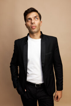Vertical portrait of stylish handsome brutal executive manager in white t-shirt and black jacket standing against brown studio background with hand in pocket, looking up with puzzled face