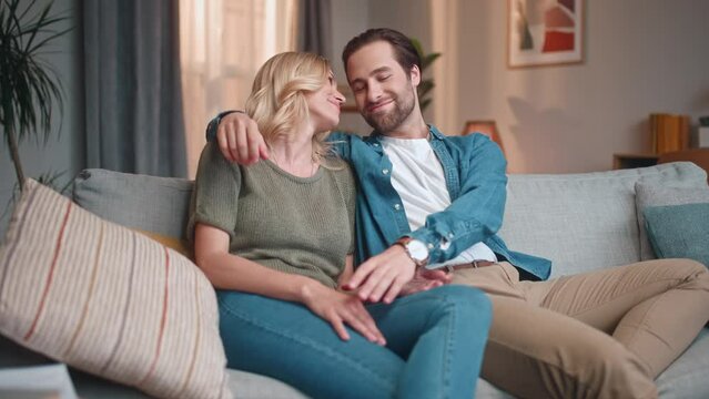 Cute husband and gorgeous wife sitting on sofa hugging and cuddling, kissing together. Lovely couple of beautiful man and woman having great time at home in living room. Love concept.