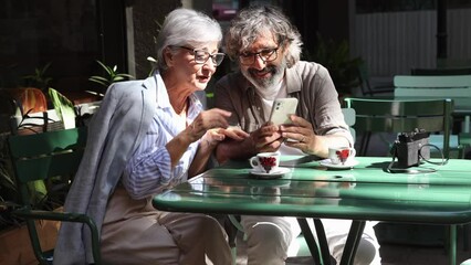 Senior couple sitting on a terrace looking at social networks on the smartphone and drinking coffee