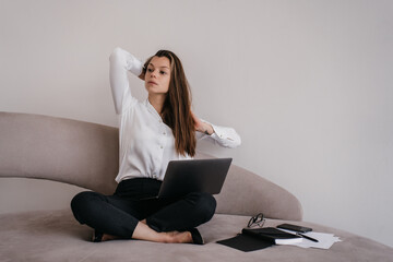 Tired brunette businesswoman with long hair sits on sofa  legs crossed using laptop looking aside stretches after hard work. Pretty overloaded hispanic girl feels fatigue. Loneliness. Working people.