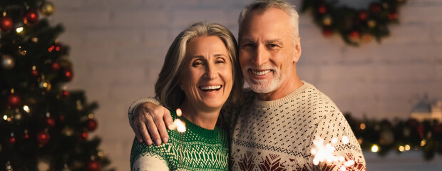 happy mature couple in holiday sweaters smiling near shiny sparklers on christmas eve, banner