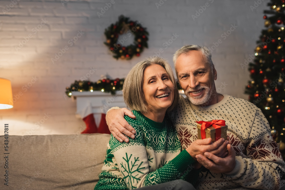 Wall mural happy middle aged man in sweater hugging cheerful wife with christmas present