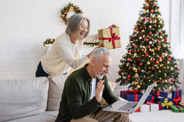 cheerful middle aged woman in glasses showing christmas present near husband during video call