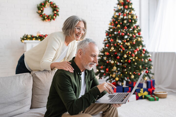 cheerful middle aged woman in glasses hugging husband during video call near christmas tree