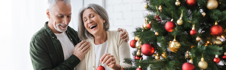 cheerful middle aged woman laughing while holding bauble near husband and christmas tree, banner