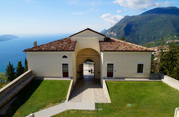 Fototapeta premium Mediterranean church Madonna di Montecastello in Tignale overlooking vast blue lake Gardasee with the Italian Alps against the clear blue sky in the background, Northern Italy, Lombardy