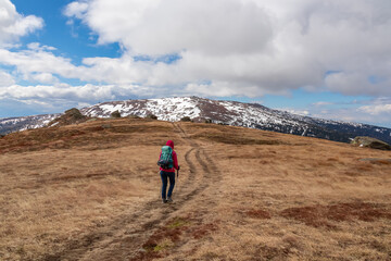 Fototapeta premium Woman with backpack hiking in cold windy winter at Ladinger Spitz in the Saualpe mountain range, Lavantal Alps, Carinthia, Austria, Europe. Hike path along on alpine pasture, clouds. Tourism Wolfsberg