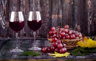 Two glasses of red wine and grapes in a straw basket, wine jug on a dark wooden background. Autumn composition with yellow and green leaves.