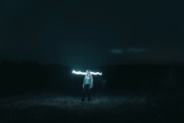 Blond girl standing in a field at night with a blue bright glow coming from behind