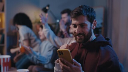 Positive bearded cheerful man looking at his smartphone with happy emotions during football game. Handsome unshaven male fan using modern smartphone on at home. Football fans concept.
