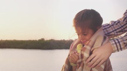 Cute girl holds fluffy yellow duckling in hands smiling on river bank against lush trees under clear sky at sunset closeup. Caring mother covers little daughter with warm plaid in summer evening - Powered by Adobe