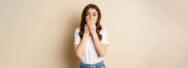 Shocked brunette woman gasping, looking speechless at camera, cover mouth with hands, standing over...