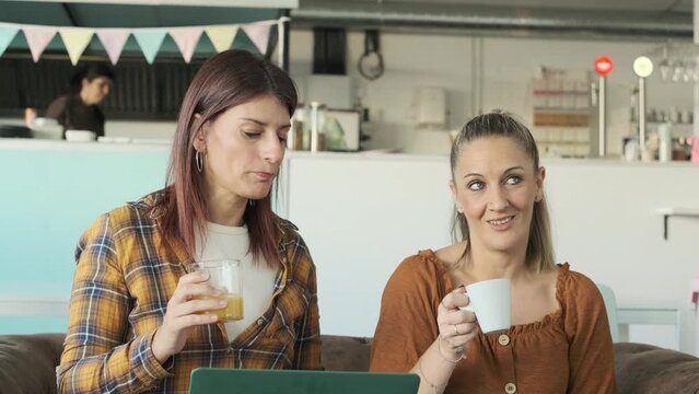 Two smiling women talking and using a laptop while drinking coffee and juice in a coffee shop.