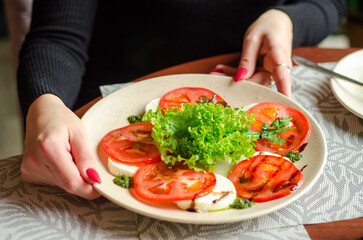 Plate of fresh salad of tomatoes, mozzarella and lettuce, on a light plate in a restaurant, female hands offer it to us