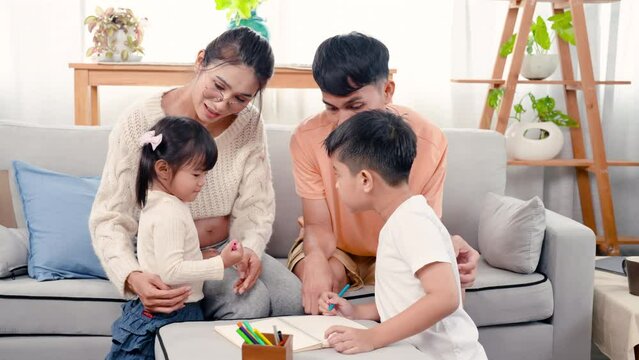 4K, family love warmth. Asian parents are lounging on sofa in  living room of house and their son and daughter drawing on coloring book with mom and dad guiding next to woman. Pregnancy, big belly.