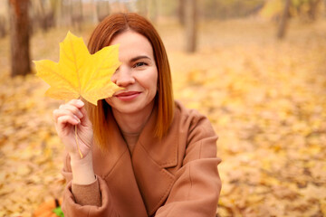 Beautiful elegant woman in autumn park holds an autumn yellow maple leaf near her face. Young woman in a beige coat walks outdoors in the park, concept autumn. High quality photo
