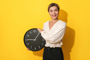 Woman with silly facial expression holding big clock on yellow background - obrazy, fototapety, plakaty