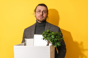 Upset man holding box with personal items after job resignation against yellow background