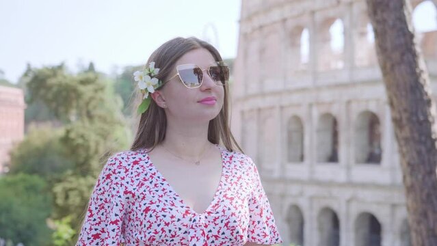 Young woman in glasses stands against large Colosseum building in Rome on sunny day. Female tourist enjoys summer vacation in Italy closeup