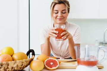 Woman drinking freshly squeezed homemade grapefruit juice in white kitchen