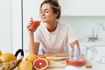 Woman drinking freshly squeezed homemade grapefruit juice in white kitchen