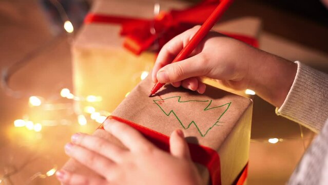Close up child hand paints and sign gift box New year present packaging. Making surprise for family. Children's paper craft school kindergarten. Christmas eve holiday party lights and ringing bells.