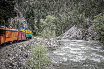 Steam engine rounding a curve with mountain cliff on one side and rushing river on other in pine...