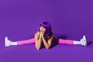 Cheerful woman wearing colorful sportswear doing splits against purple background