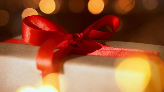 Close up Christmas gift craft paper box with red ribbon bow over dark lights bokeh background. New year eve holiday composition with present handmade box. Eve preparation. Family time, cozy home