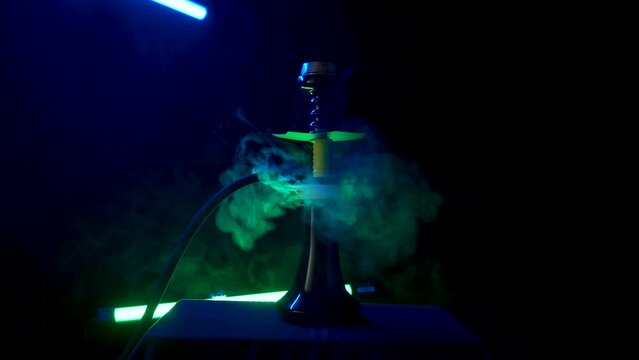 Stylish hookah with yellow shaft stands on dark table. White thick clouds of smoke rise from hot coals dissipating around hookah. Smoking hookah in bar