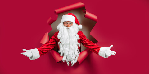Santa Clause peeking through hole on red paper while gesturing hands like welcome. New Year advert