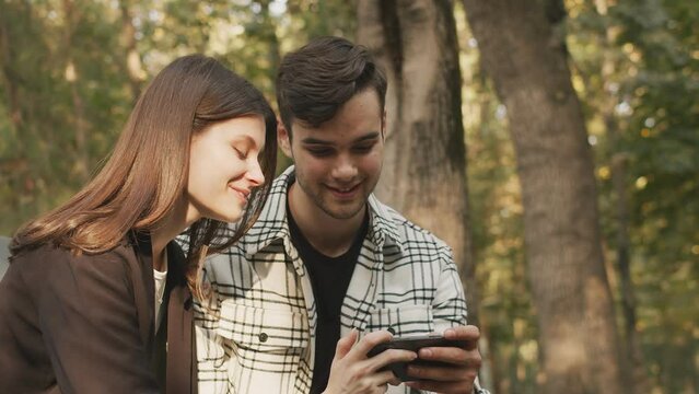 Girlfriend and boyfriend watching video on a mobile handheld device while talking, green trees, nature in the background. Using smartphone together, showing pictures, sharing memories