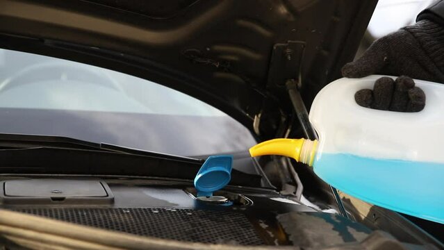 Close-up detail of driver or mechanic hand in gloves pouring blue antifreeze liquid for winter car windshield screen washing with bootle watering can. Car seasonal service and maintenance concept