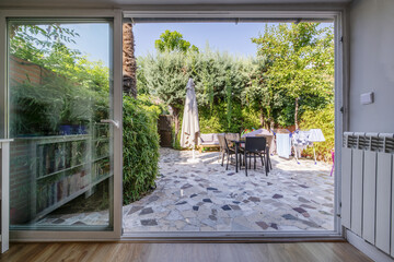 Large white aluminum and glass window open in a living room to give access to a terrace with cement and stone floors and hedges and trees