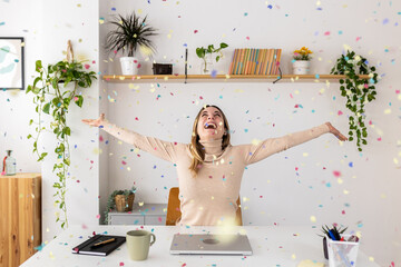 Happy young businesswoman celebrating success throwing confetti overhead sitting at desk in home...