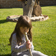 Image of an adorable little brunette girl sitting on a green grass while learning to play the flute  © gianni