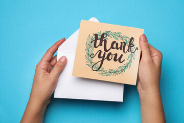 Woman holding envelope and card with phrase Thank you on light blue background, top view