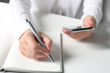 Woman with smartphone writing in notebook at white table in office, closeup