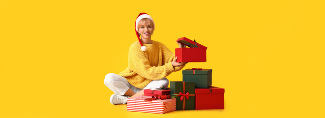 Beautiful young woman in Santa hat and with many Christmas gifts on yellow background