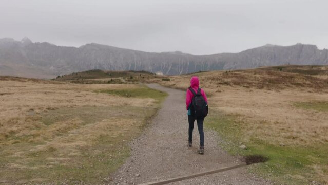 A girl in a deep pink jacket walking on a path in Aple de Siusi in a cloudy day.Wide shot. South Tirol, Italy.