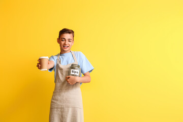 Male barista with cup of coffee and tip jar on yellow background