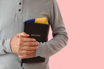 Mature man with Holy Bible and flag of Ukraine praying on pink background, closeup