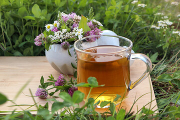 Cup of aromatic herbal tea, pestle and ceramic mortar with different wildflowers on green grass outdoors